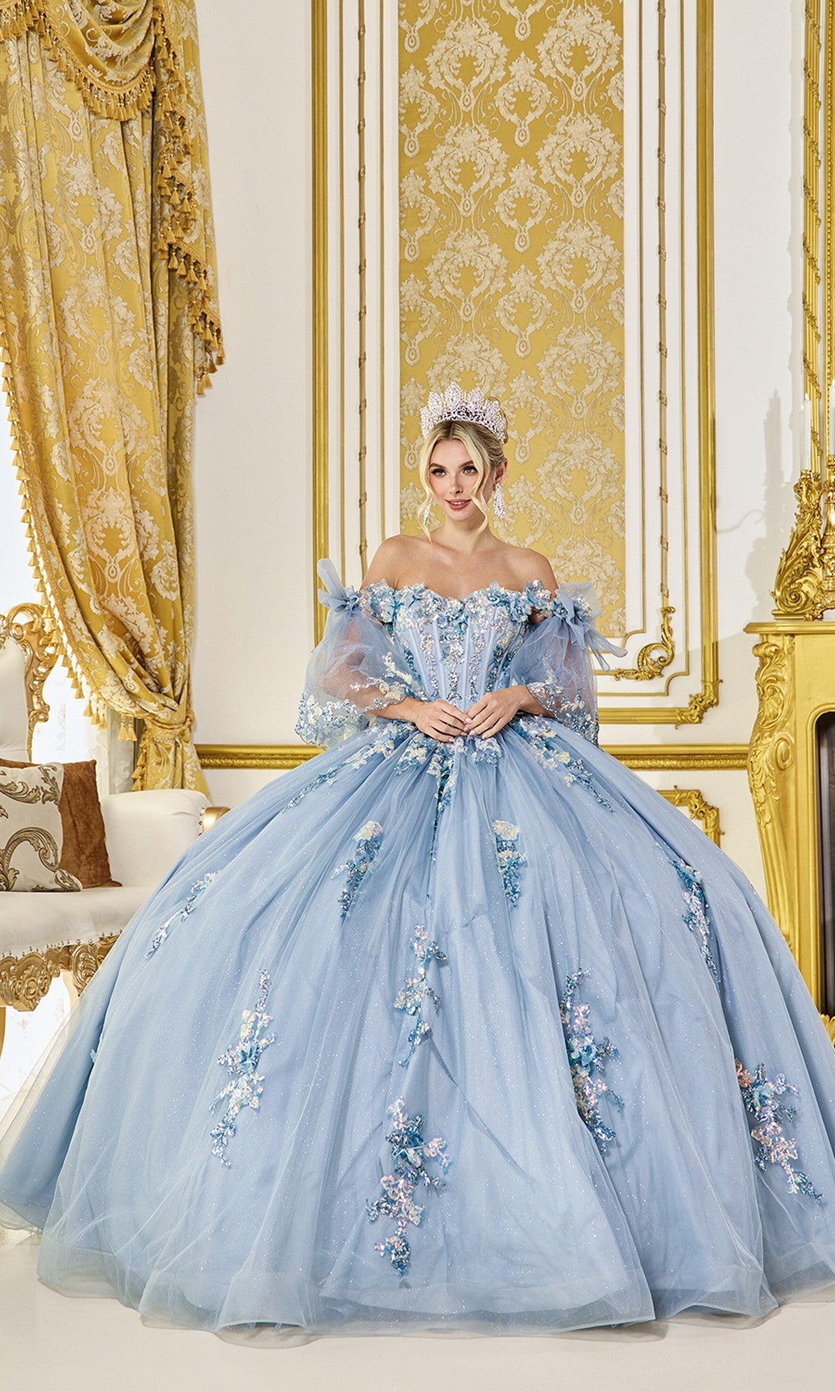 A woman wears the Quinceanera Dress 5072 By Dancing Queen in dusty blue with floral details and a jeweled crown, standing in an opulent gold-decorated room.